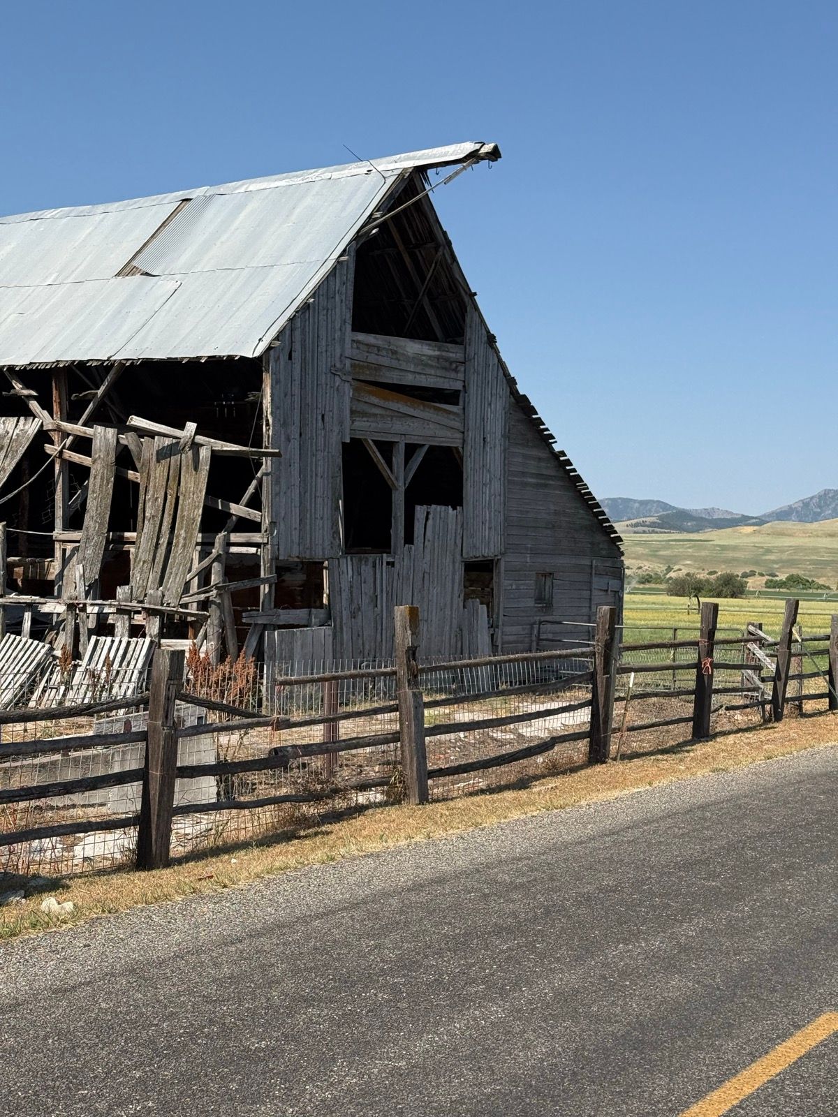 An old, weathered barn with a metal roof, partially collapsed, is situated next to a dirt road in Nibley, Utah. A wooden fence lines the foreground, and the landscape features rolling hills and a clear blue sky in the background.
