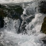 A close-up of rushing water in a stream, with bubbles and waves creating a dynamic, frothy appearance. The water flows over rocks, surrounded by green moss, in Paradise, Utah.