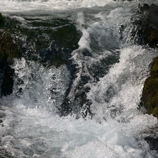 A close-up of rushing water in a stream, with bubbles and waves creating a dynamic, frothy appearance. The water flows over rocks, surrounded by green moss, in Paradise, Utah.