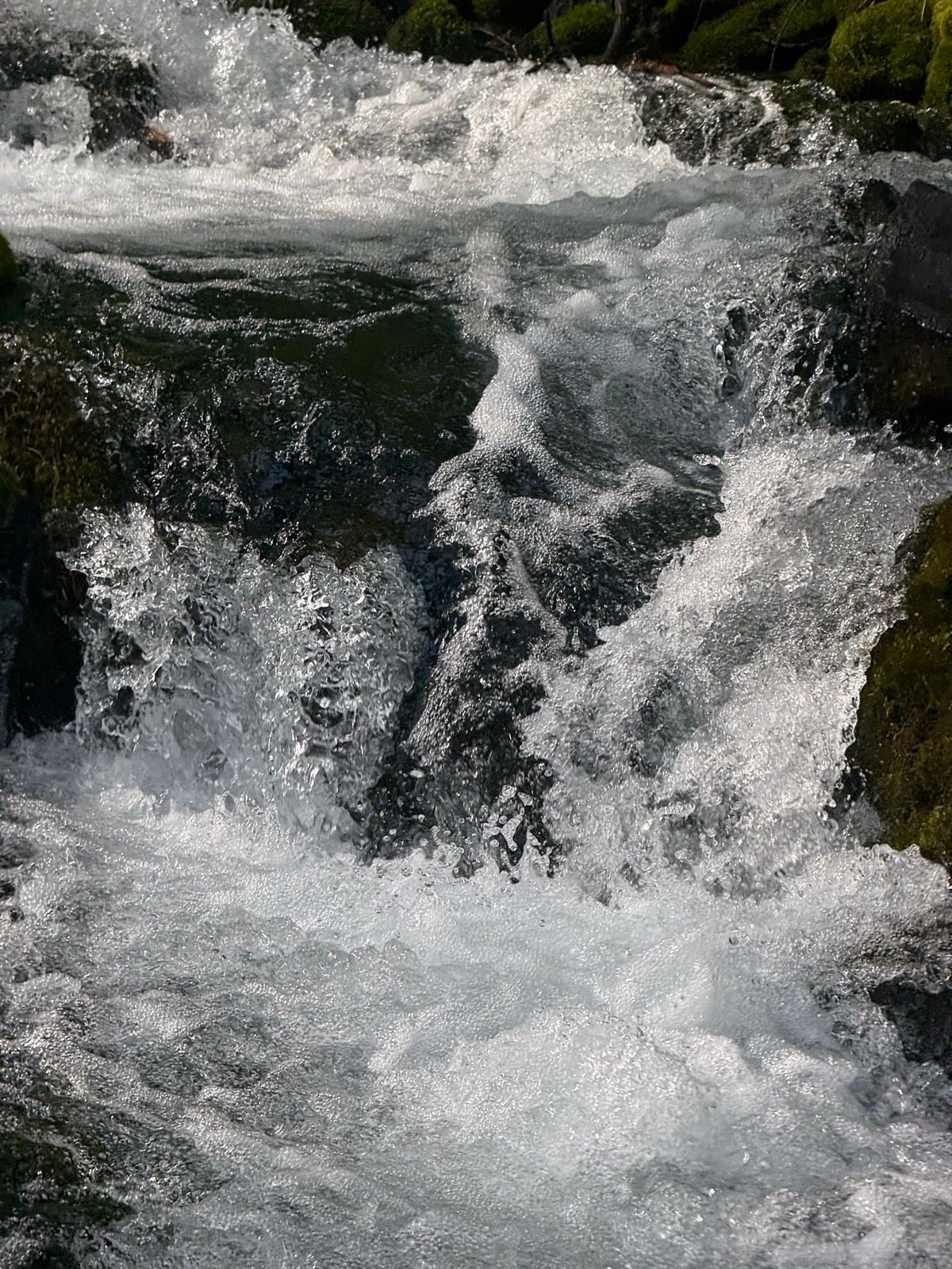 A close-up of rushing water in a stream, with bubbles and waves creating a dynamic, frothy appearance. The water flows over rocks, surrounded by green moss, in Paradise, Utah.