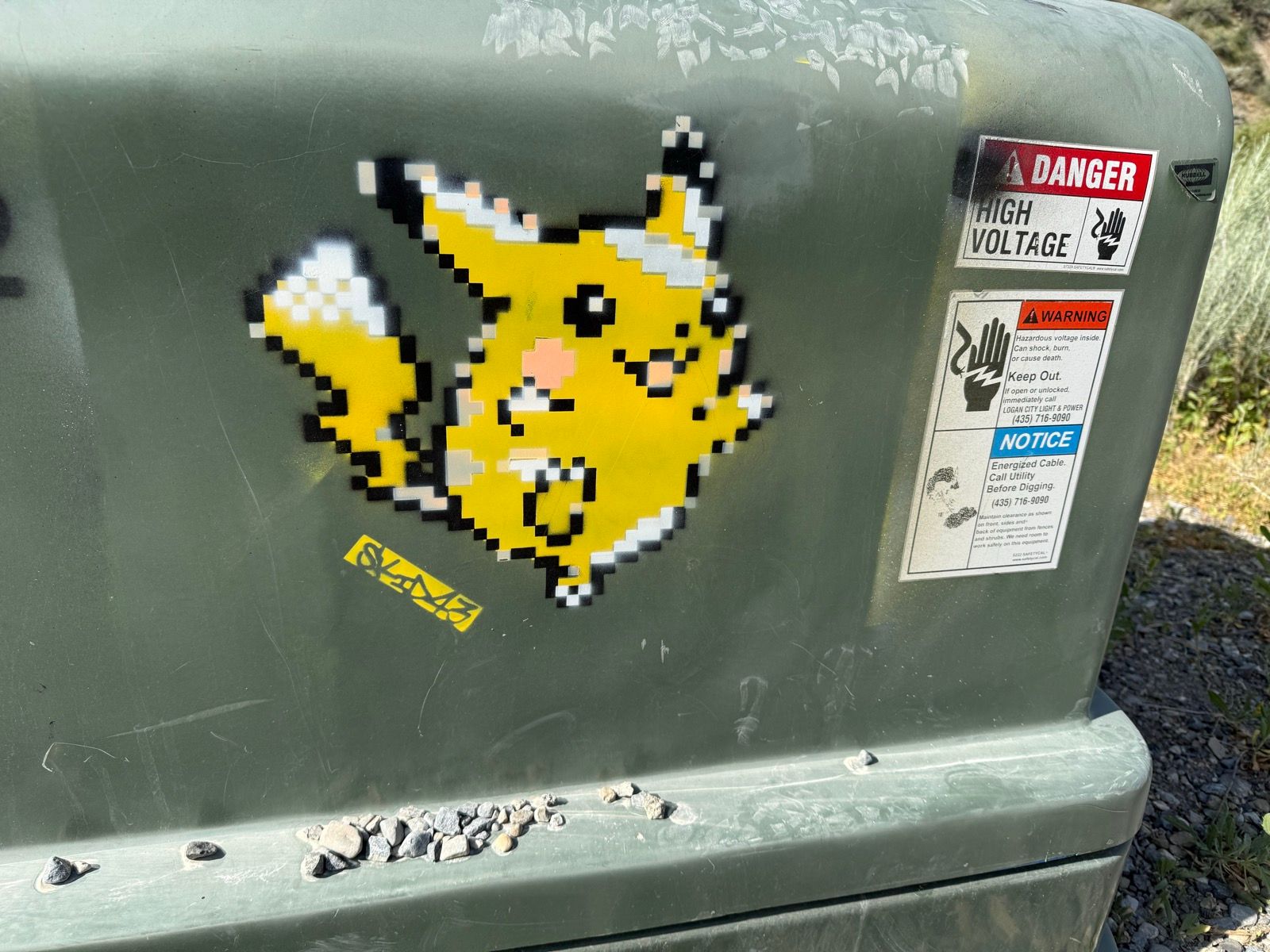 A pixelated yellow cartoon character sticker is attached to a green electrical box, which also has warning labels about high voltage and hazardous conditions. Small rocks are scattered at the bottom of the box. The photo was taken in Paradise, Utah.