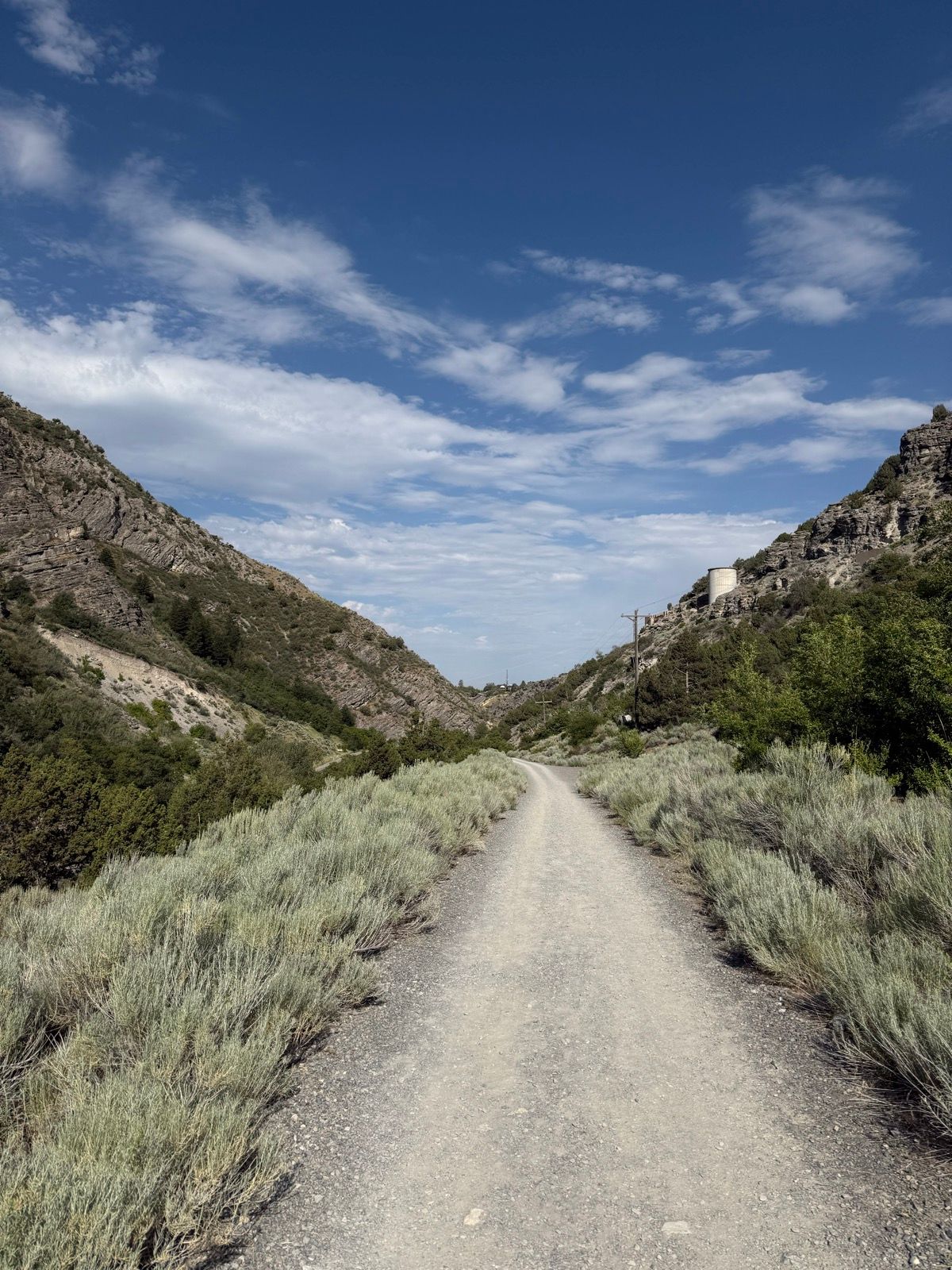 A dirt path winds through a canyon in Paradise, Utah, surrounded by green shrubs and rocky hills under a blue sky with scattered clouds.