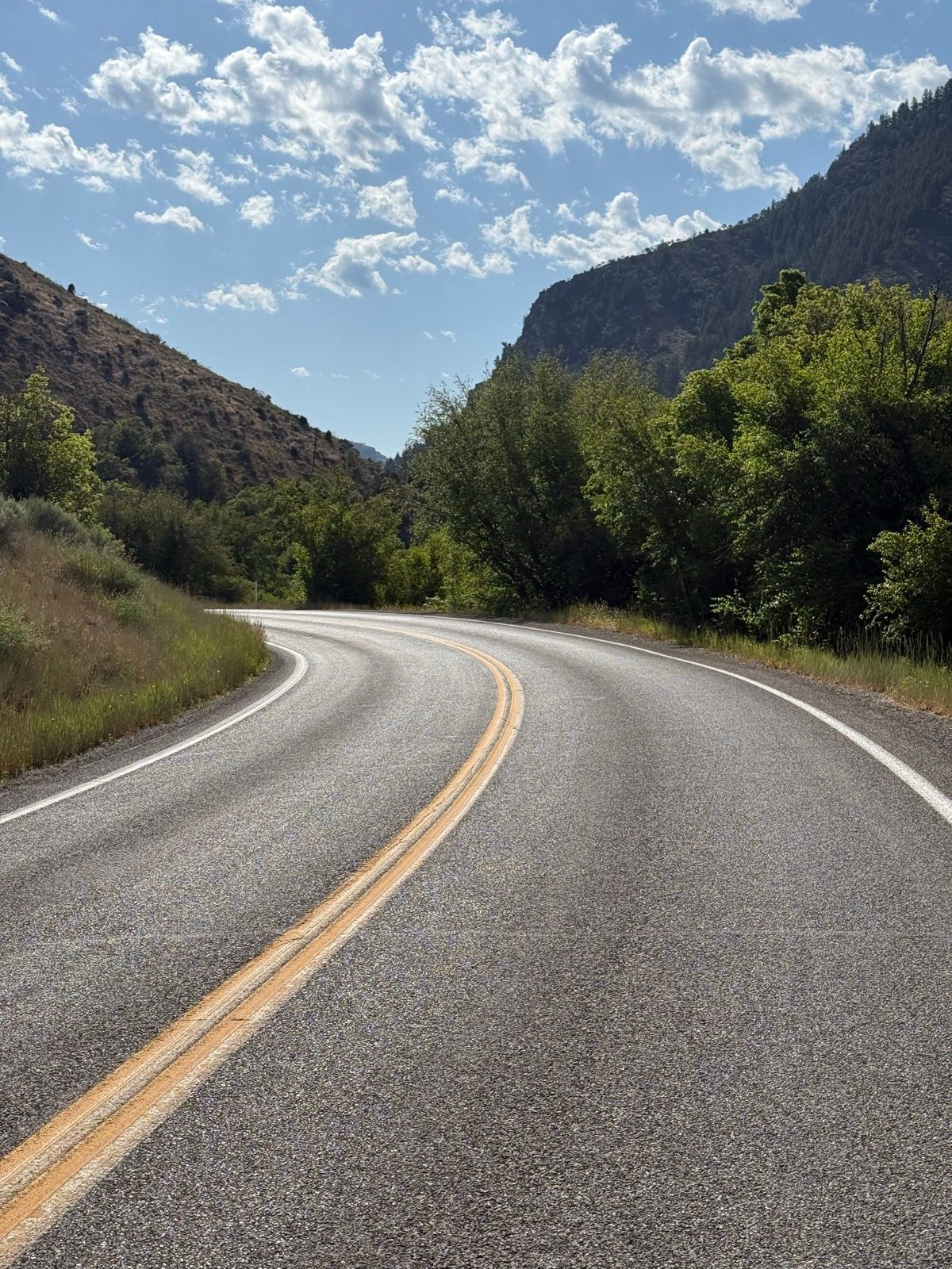 A curving road bordered by green trees and rocky hills under a partly cloudy sky in Nibley, Utah.
