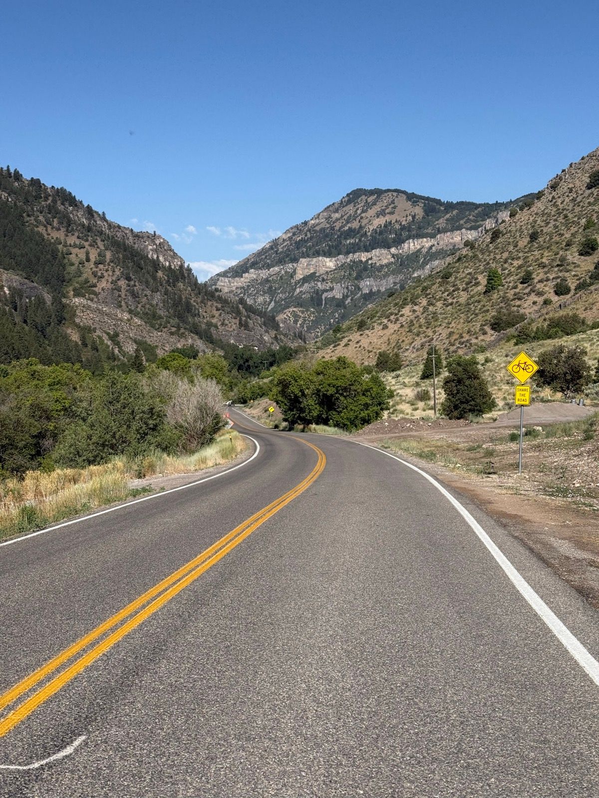A winding road surrounded by mountains and trees in Nibley, Utah, under a clear blue sky. A bicycle warning sign is visible along the roadside.