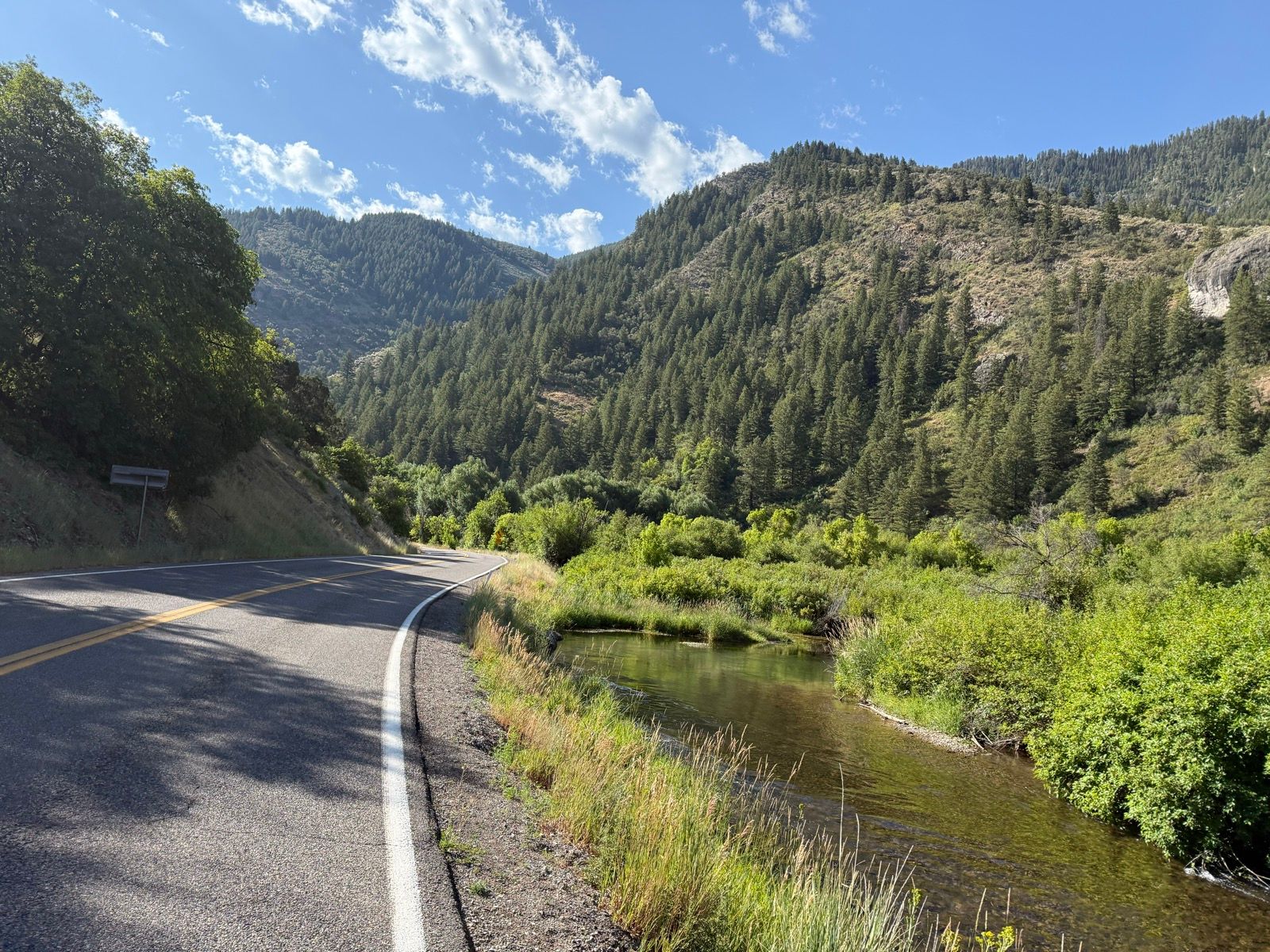 A winding road curves alongside a serene stream, surrounded by lush greenery and tall pine trees. In the background, rolling hills are visible under a partly cloudy blue sky. This scene is set in Nibley, Utah.