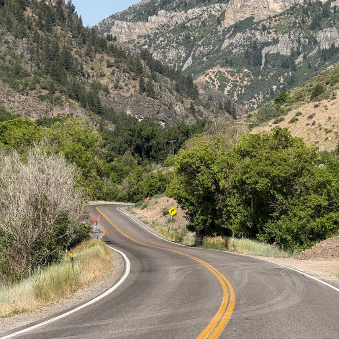 A winding road curves through a mountainous landscape in Nibley, Utah. Lush greenery lines the road, with rocky hills and steep slopes in the background. The sky is clear and blue, and construction signs are visible along the roadside.