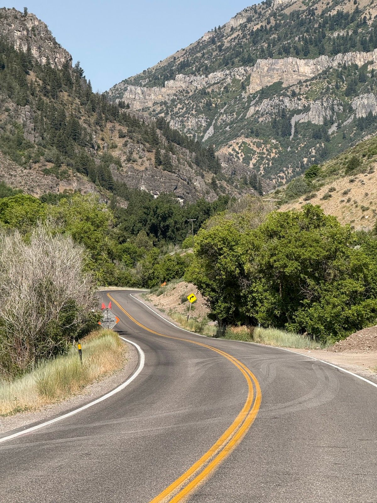 A winding road curves through a mountainous landscape in Nibley, Utah. Lush greenery lines the road, with rocky hills and steep slopes in the background. The sky is clear and blue, and construction signs are visible along the roadside.