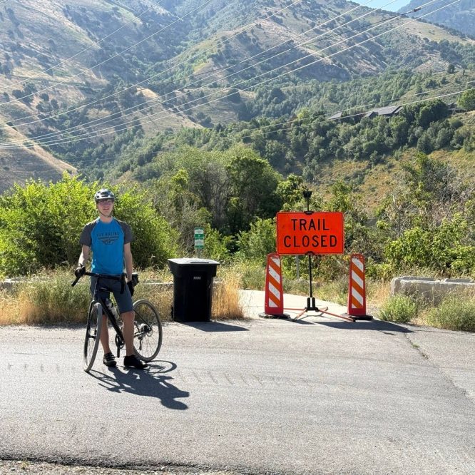 A person in a blue t-shirt and helmet stands next to a bicycle at a road intersection. There is a bright orange "TRAIL CLOSED" sign ahead, indicating that access is restricted. Lush green hills and mountains are visible in the background on a sunny day.