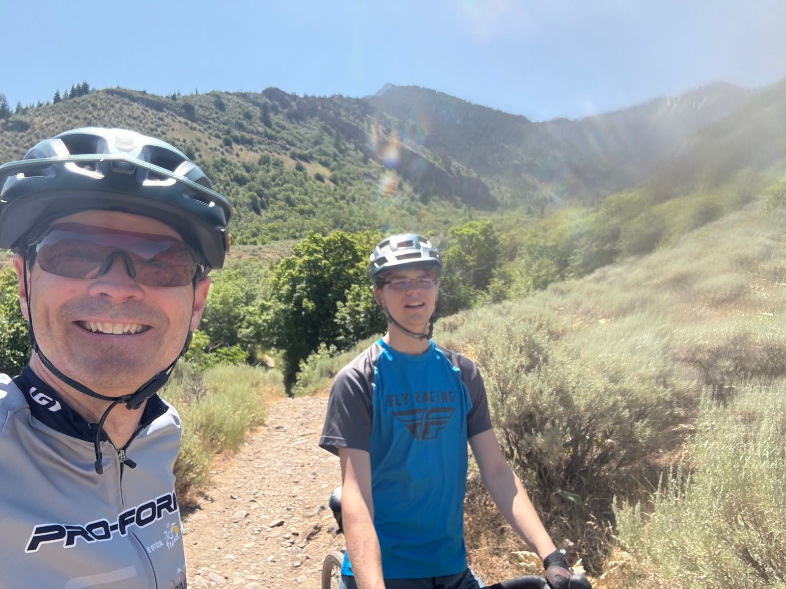 A man and a boy are posing for a selfie while mountain biking on a rocky path in Providence, Utah. They are both wearing helmets and sunglasses, with green hills and trees in the background under a clear blue sky.