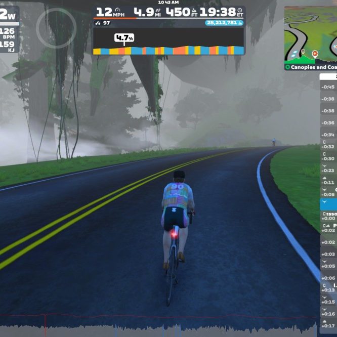 A cyclist in a virtual environment on Zwift rides along a road surrounded by misty trees. The screen displays various performance stats, including power, speed, and distance. Additionally, a list of nearby riders and their performance metrics is shown on the right side of the screen.