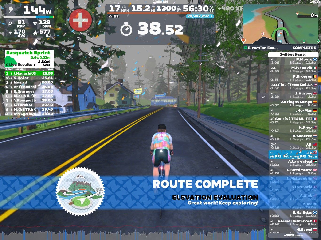 A screenshot from Zwift shows a virtual cycling environment with a cyclist in the foreground riding on a road. The cyclist is wearing a colorful jersey and is about to complete the