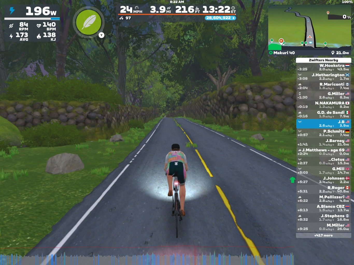A cyclist in a virtual environment rides along a winding road surrounded by greenery. The cyclist is equipped with a smart trainer, displaying performance metrics such as power output, speed, and elevation. A side panel shows nearby virtual riders, their stats, and a map of the route.