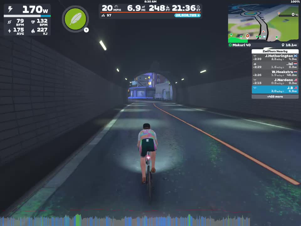 A cyclist rides through a virtual tunnel in Zwift, with a display showing power output, speed, distance, and heart rate. The interface includes a map and nearby riders' stats. The environment is illuminated with soft lighting.