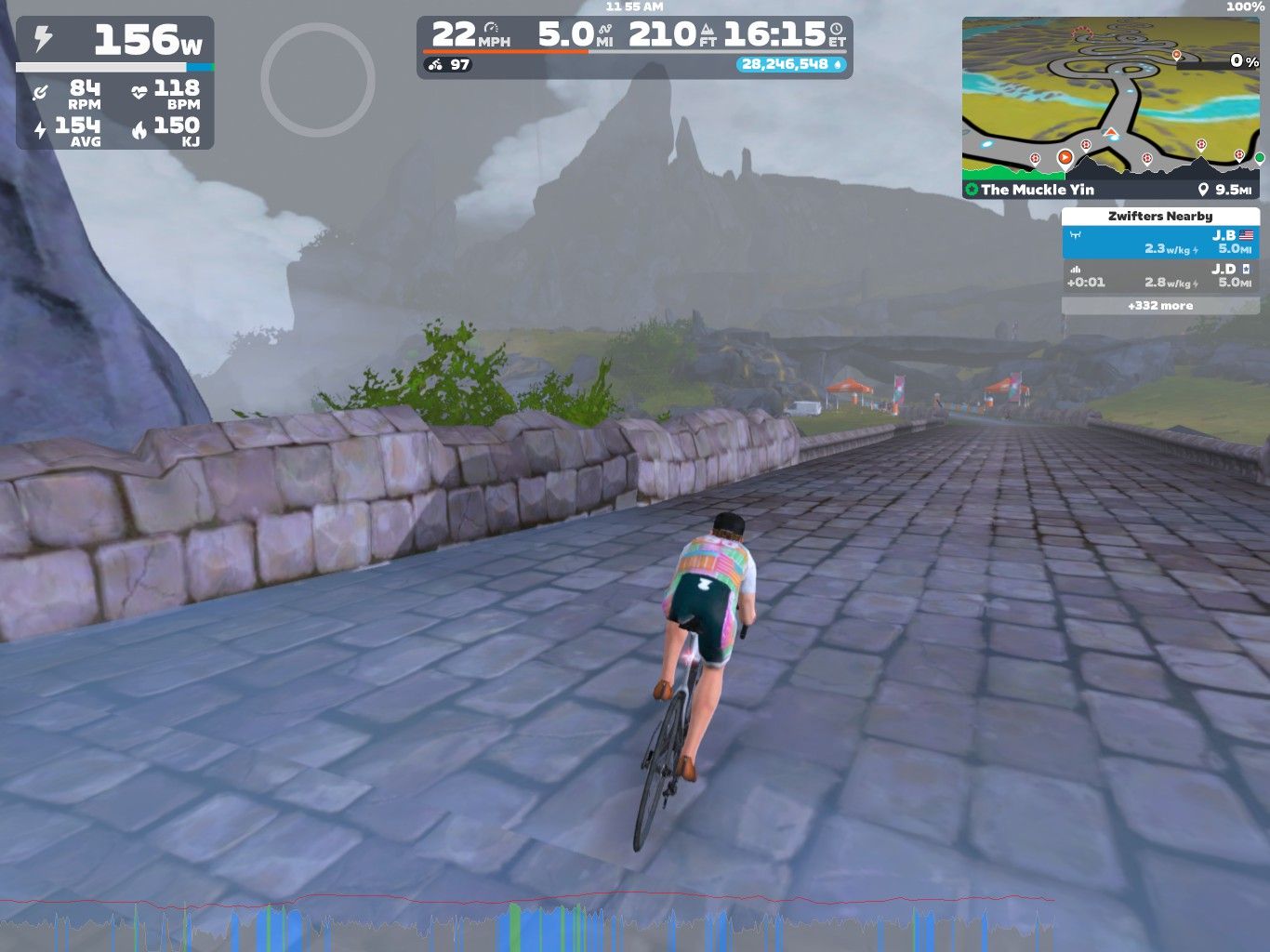 A cyclist rides on a cobblestone path in a virtual landscape, surrounded by rocky hills and fog. The screen displays various stats, including power output, speed, distance, and heart rate. The route indicated shows the cyclist's position within the Zwift virtual cycling platform.