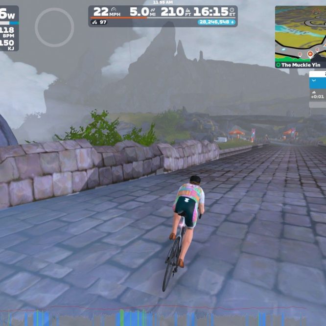 A cyclist in a colorful jersey rides on a cobblestone path in a virtual landscape, with mountains and a misty sky in the background. The display shows various cycling metrics, including speed, power, and elevation. Nearby are virtual flags and a winding road on the map.