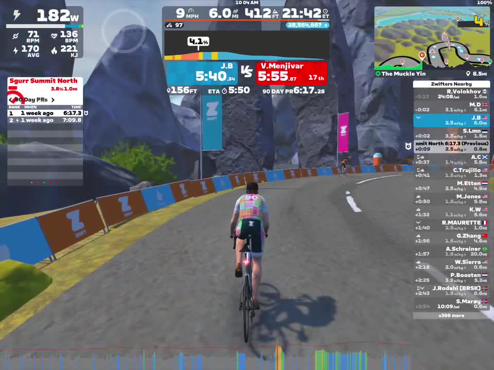 A cyclist is riding on a virtual road in Zwift, surrounded by digital mountains and a colorful barrier. The screen displays data such as power output, speed, distance, and the cyclist's position in a race. Other names and statistics of nearby riders are visible on the right side.