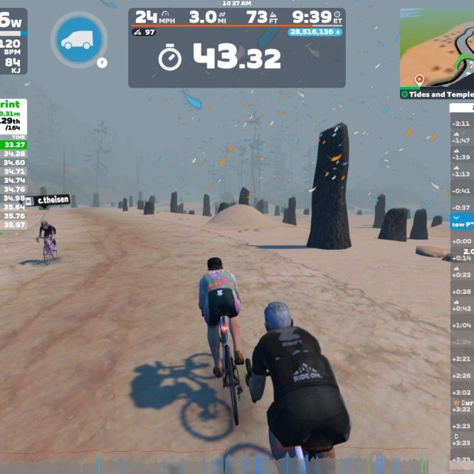 A digital cycling scene in Zwift shows two cyclists competing in a sprint challenge. The terrain features a sandy path with scattered stone formations. On the left, live results display rankings and times for participants. The dashboard indicates the cyclists' speed, power output, and other stats. A foggy atmosphere adds to the virtual environment.
