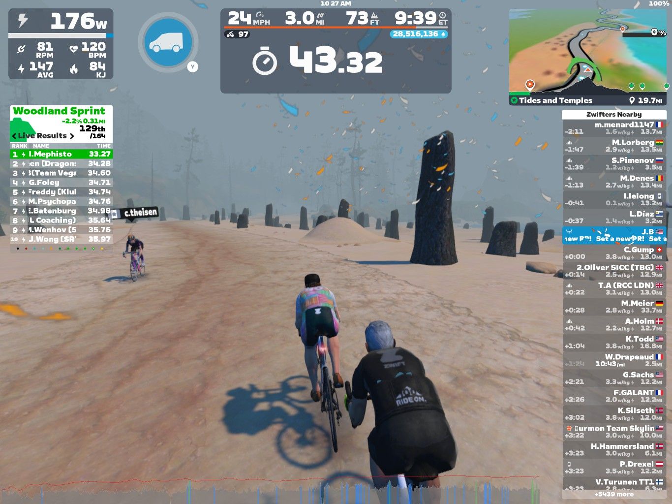 A digital cycling scene in Zwift shows two cyclists competing in a sprint challenge. The terrain features a sandy path with scattered stone formations. On the left, live results display rankings and times for participants. The dashboard indicates the cyclists' speed, power output, and other stats. A foggy atmosphere adds to the virtual environment.