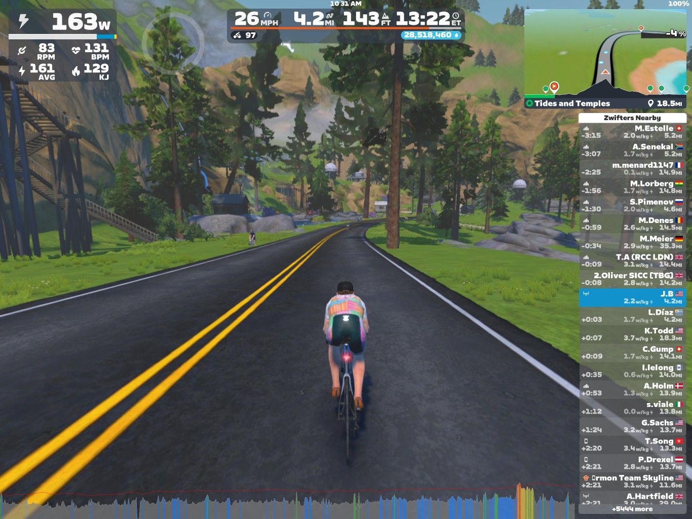 A cyclist riding on a virtual road in a scenic landscape, surrounded by trees and mountains. The screen displays cycling statistics, including power output, speed, and heart rate, along with a list of nearby virtual riders.