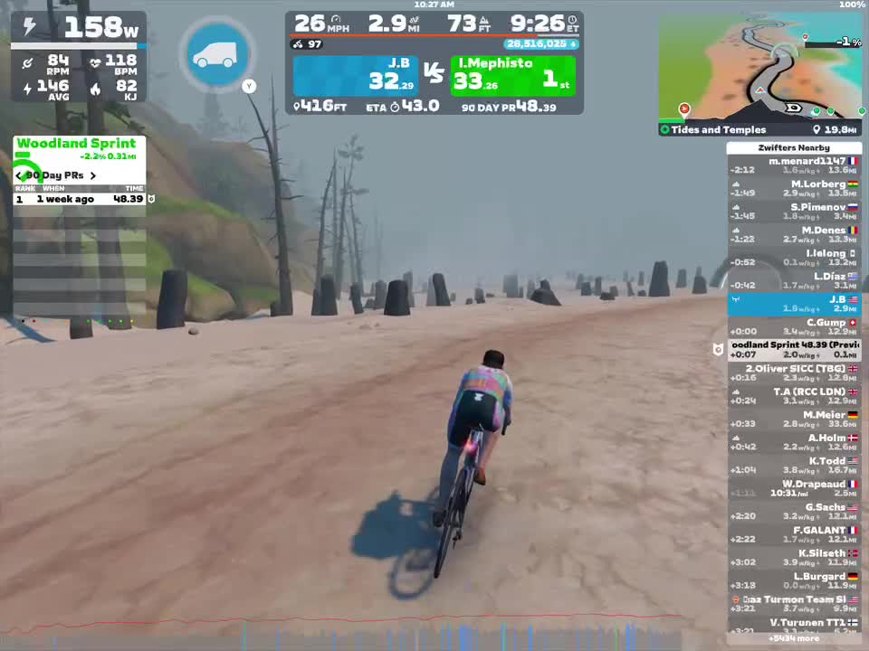 A cyclist is riding on a dirt path in a virtual environment, surrounded by rocky terrain and trees. On the screen, various cycling statistics and leaderboards are displayed, including speed, power output, and positions of nearby riders. The setting appears to be part of a gaming simulation.