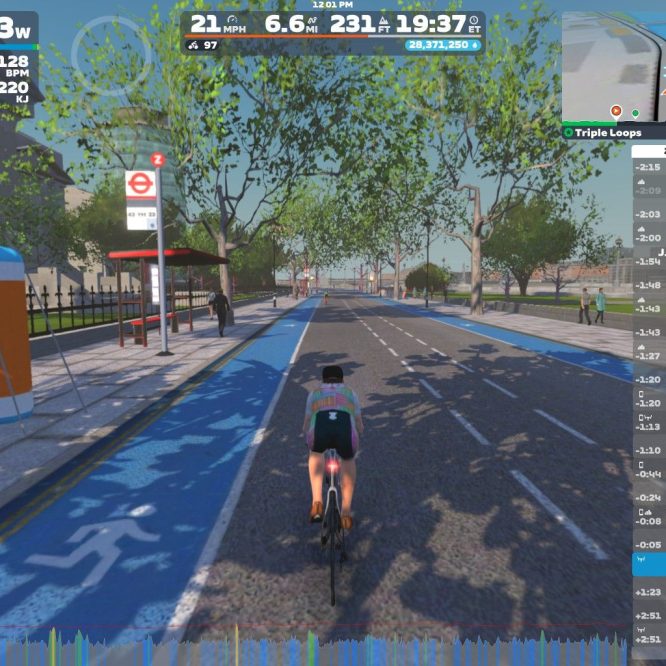 A cyclist rides along a blue bike lane in a virtual environment, with trees lining the road. There is a bus stop nearby, and a few pedestrians are visible in the background. The cyclist is wearing a colorful jersey, and a statistics panel shows performance data such as speed and power.