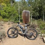 A mountain bike is standing next to a wooden sign that reads "Mount Naomi Wilderness" in Green Canyon, Utah. The background features green trees and shrubs in a natural setting.