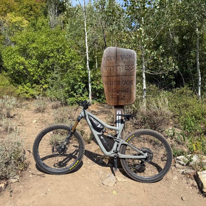 A mountain bike is standing next to a wooden sign that reads "Mount Naomi Wilderness" in Green Canyon, Utah. The background features green trees and shrubs in a natural setting.