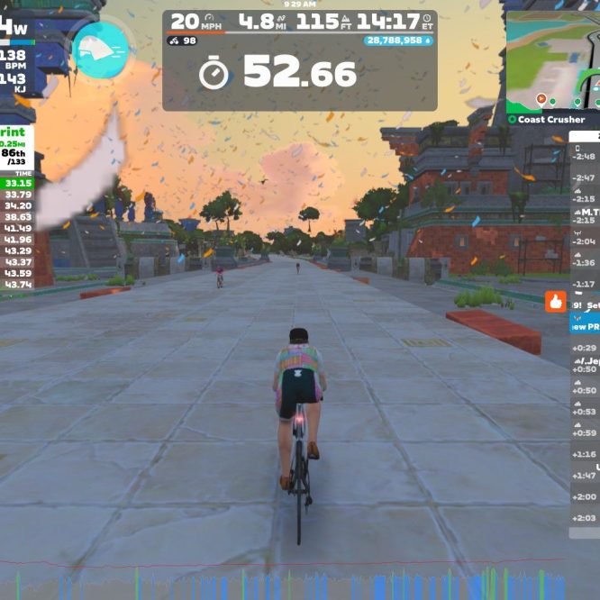 A cyclist participates in a virtual race on the Zwift platform, displaying stats on power, speed, and heart rate. The screen shows the current race results, nearby competitors, and the cyclist's progress along the virtual course. The environment features a colorful landscape with stylized buildings and trees in the background.