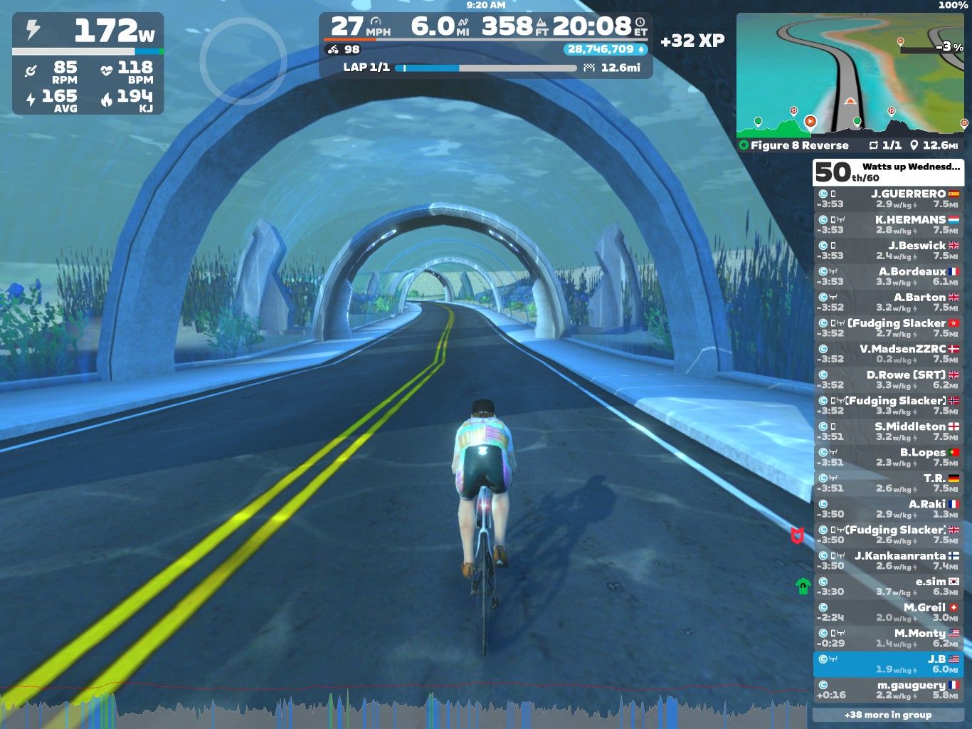 A cyclist rides on a virtual road in Zwift, surrounded by underwater-themed arches and aquatic plants. The screen displays cycling stats, including speed, distance, and power output, along with a leaderboard on the right side.