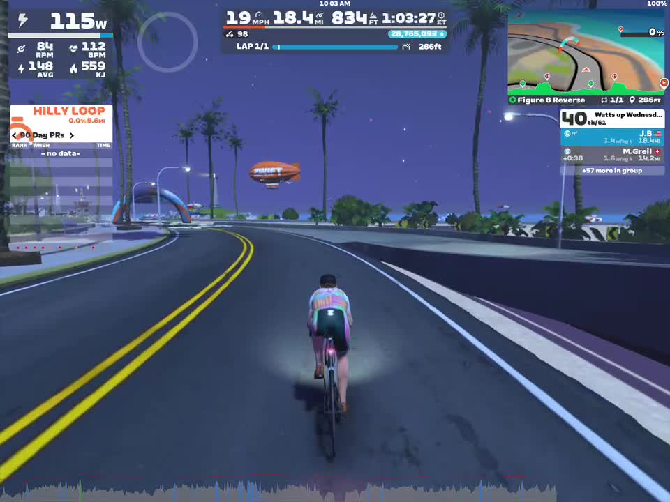A cyclist rides along a virtual road in a simulated environment, surrounded by palm trees and an evening sky. The screen displays various metrics including speed, RPM, and heart rate, along with a mini-map and additional rider information. A blimp can be seen in the background, and the course is labeled as 