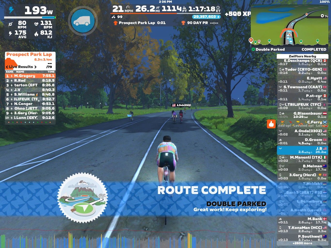 A screenshot from the Zwift cycling app showing a virtual cyclist finishing a lap. The interface displays performance metrics, including power output and rank. A list of live results shows participant names and lap times, while a banner at the bottom indicates that the route is complete. Lush trees line the road, creating a scenic backdrop for the ride.