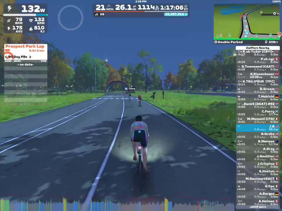A cyclist rides on a virtual road in Zwift during an evening scene. The cyclist is traveling at a speed of 21 mph and has a power output of 132 watts. Nearby cyclists are visible, and a tool panel displays performance stats and a map of the area. The setting shows trees and greenery along the sides of the road.