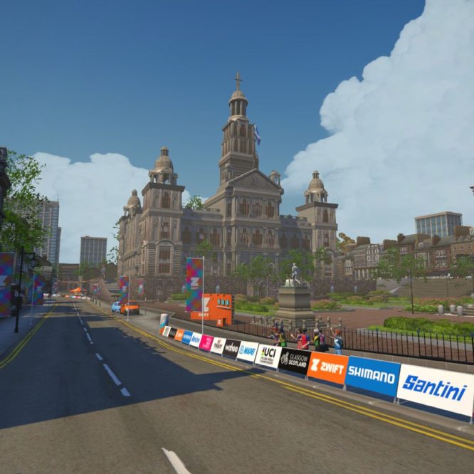 A virtual street scene from Zwift showing a wide road lined with colorful banners and signage. In the background, a large historic building with domes is visible under a partly cloudy blue sky. People can be seen along the road, cheering, while trees and a statue add to the park-like setting.