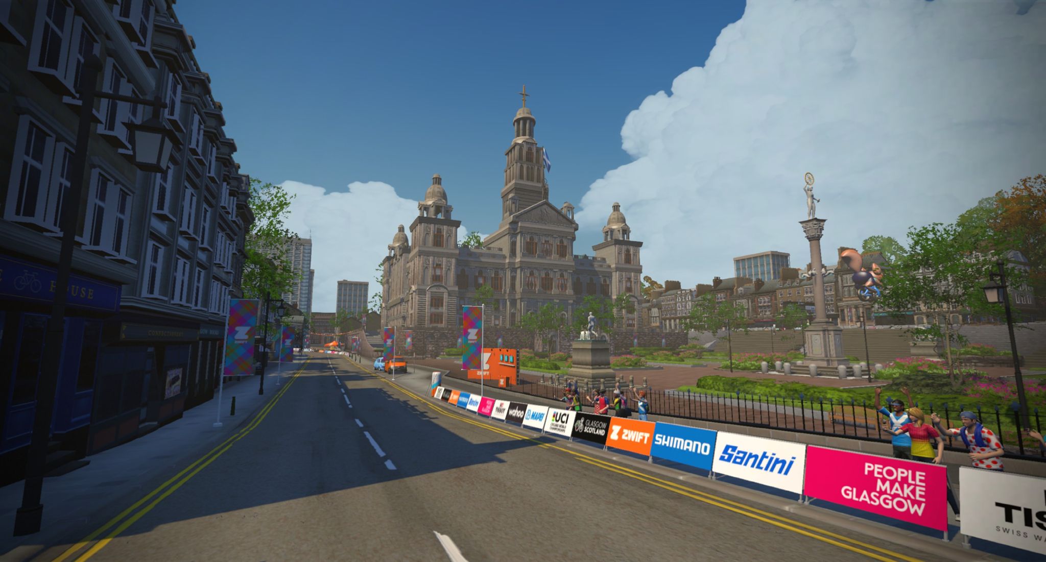 A virtual street scene from Zwift showing a wide road lined with colorful banners and signage. In the background, a large historic building with domes is visible under a partly cloudy blue sky. People can be seen along the road, cheering, while trees and a statue add to the park-like setting.