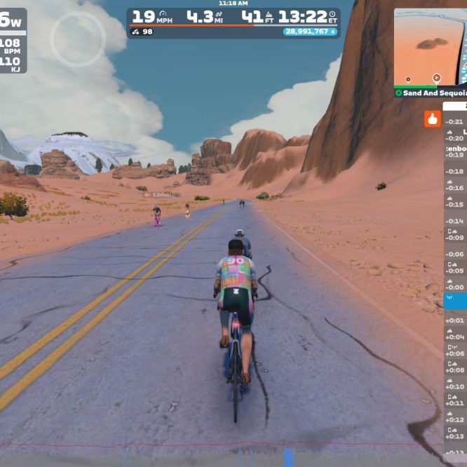 A virtual cycling scene in Zwift showing a cyclist on a road with mountains and rocky terrain in the background. The cyclist is wearing a colorful outfit and is accompanied by other riders. The on-screen stats display power, speed, heart rate, and various nearby users.