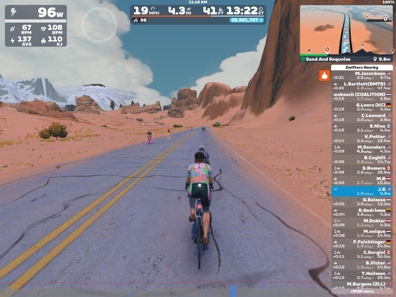 A virtual cycling scene in Zwift showing a cyclist on a road with mountains and rocky terrain in the background. The cyclist is wearing a colorful outfit and is accompanied by other riders. The on-screen stats display power, speed, heart rate, and various nearby users.