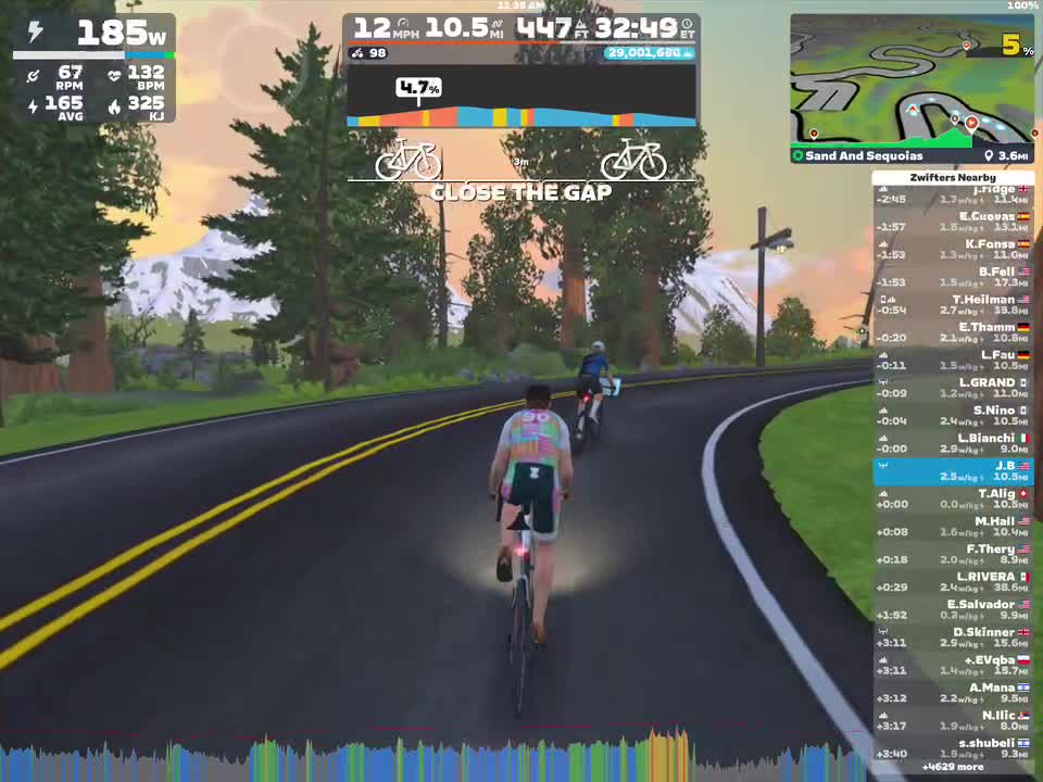 A virtual cycling scene in Zwift shows two cyclists riding on a winding road surrounded by trees and mountains. The screen displays important cycling metrics such as wattage, speed, and heart rate, along with a leaderboard of nearby riders. The sunset casts a warm glow over the landscape.