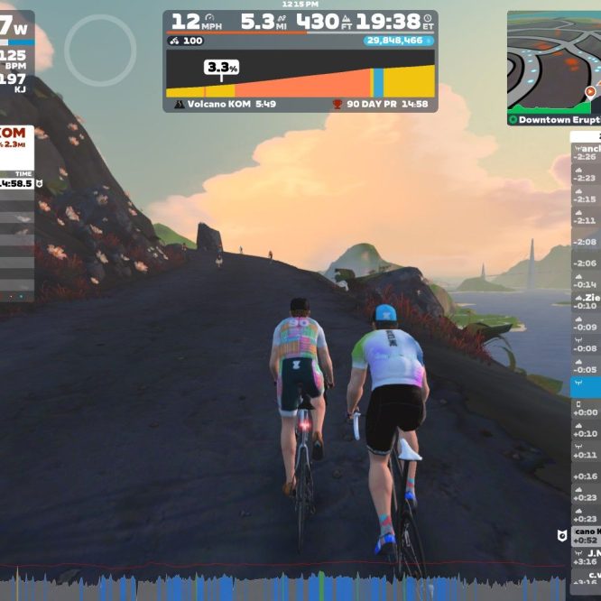 Two cyclists are riding on a virtual road in Zwift, with a scenic background featuring hills and a colorful sky. The cyclists are approximately shoulder to shoulder, and various on-screen stats display their speed, power output, and other performance metrics. The setting includes a view of a mountain labeled "Volcano KOM."
