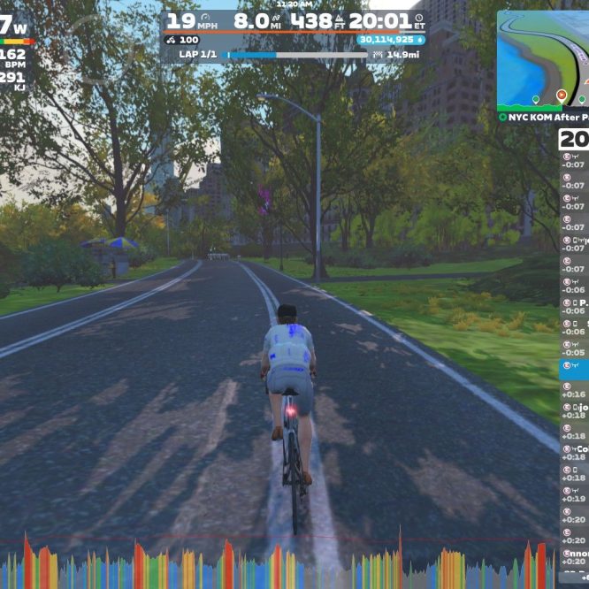 A cyclist in a blue and white jersey rides on a well-maintained path surrounded by trees in a virtual cycling environment. The screen displays various statistics, including power output, speed, and heart rate, along with a map and leaderboard on the side. The setting appears to simulate a sunny day, enhancing the cycling experience.