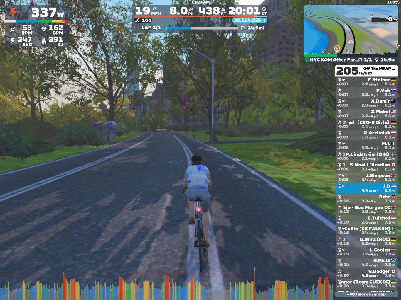 A cyclist in a blue and white jersey rides on a well-maintained path surrounded by trees in a virtual cycling environment. The screen displays various statistics, including power output, speed, and heart rate, along with a map and leaderboard on the side. The setting appears to simulate a sunny day, enhancing the cycling experience.