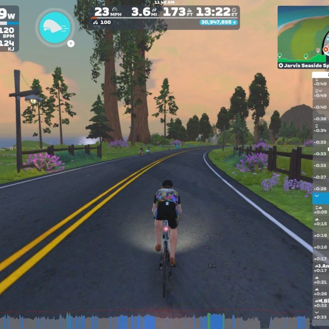 A cyclist rides on a virtual road surrounded by greenery and trees in Zwift, showcasing stats like power, speed, and distance on the screen. In the background, other cyclists can be seen along the road. The scene is set during a colorful sunrise or sunset.