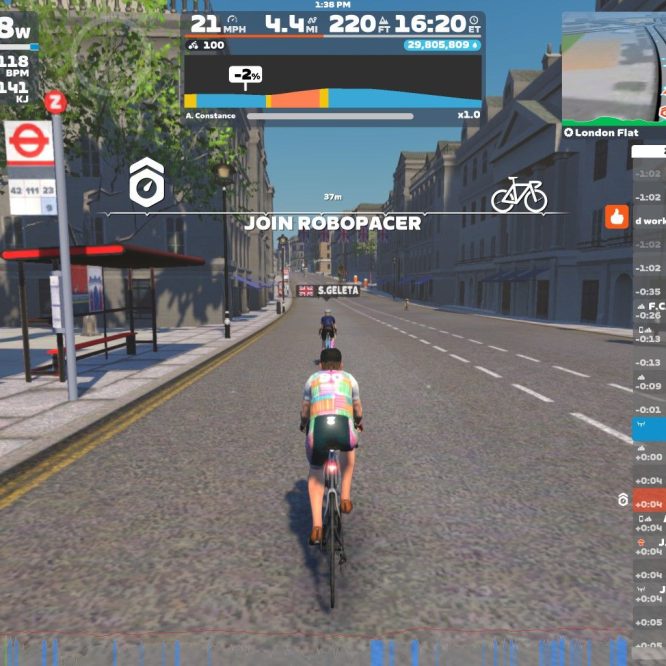 A cyclist riding on a virtual street in Zwift, with a bus stop nearby. The screen displays real-time stats such as wattage, speed, distance, and heart rate, along with a sidebar showing other users and their performance metrics. Bright sunlight and trees line the street, creating a lively outdoor atmosphere.