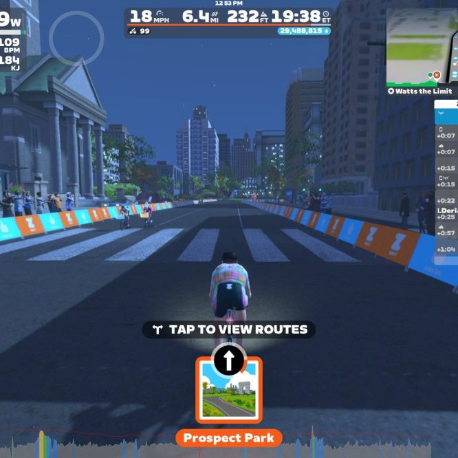 A cyclist in a virtual environment is riding down a city street in Zwift. The scene features tall buildings, a crowd of spectators, and a progress bar displaying cycling stats, including wattage, speed, and distance. The current route is labeled "Prospect Park."