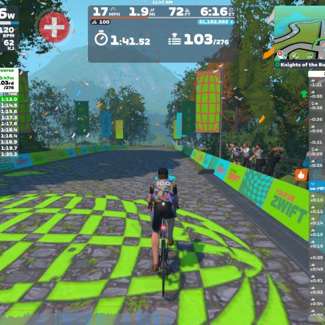 A cyclist rides on a virtual road in Zwift, surrounded by trees and colorful banners. The screen displays the rider's statistics, including power output, speed, and heart rate, along with a leaderboard showing the rankings and times of other riders in a sprint event. The scenic background features mountains and a clear sky, while confetti falls in celebration.