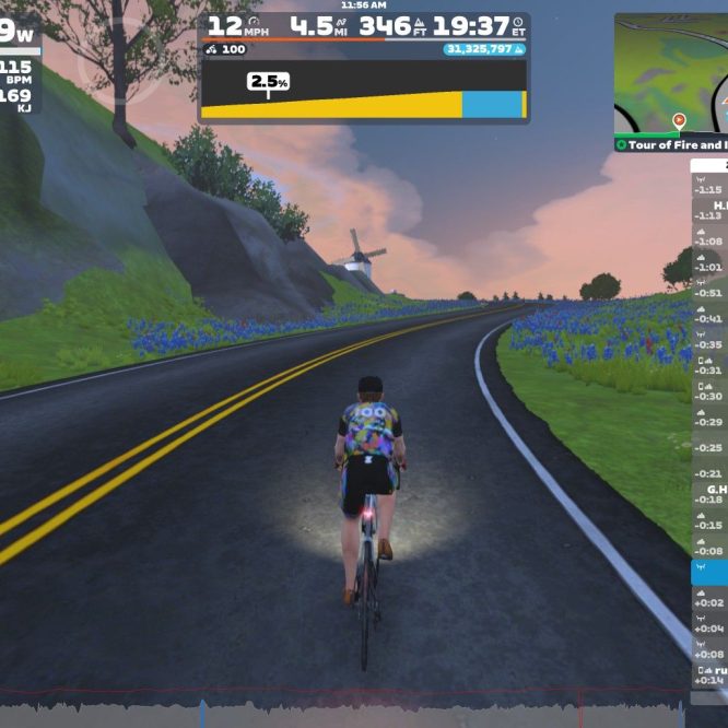 A cyclist rides on a virtual road in Zwift, surrounded by green hills and purple flowers. The cyclist has a colorful outfit and the screen displays performance metrics, including speed, distance, and heart rate. The sky in the background shows a sunset. A leaderboard on the right lists other riders nearby.