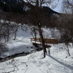 A wooden bridge crosses a small creek in a snowy landscape at Providence Canyon, Utah. Surrounding the bridge are bare trees and snow-covered ground, with mountains visible in the background under a partly cloudy sky.