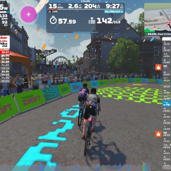A group of cyclists participating in the "Tour de Zwift" race is seen on a virtual roadway. Two cyclists are prominently featured in the foreground, riding side by side. In the background, there are cheering spectators and festive decorations. Various metrics, including power output and speed, are displayed on the screen. The event is set in a colorful, animated cityscape.