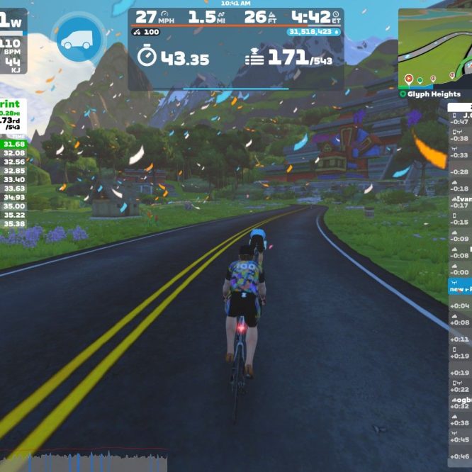 A cyclist rides on a virtual road in Zwift, with greenery and mountains in the background. The screen displays performance stats, including speed (27 mph), power (141 W), and heart rate (110 bpm), alongside race results and nearby competitors. Confetti floats in the air, indicating a festive atmosphere, while the route features yellow center dividing lines.