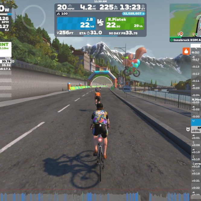 A digital cycling scene in Zwift featuring cyclists riding on a virtual road. Key metrics such as power (190W) and speed (20 mph) are displayed on the screen, along with race statistics and rider rankings. A colorful banner and bike-themed decorations are visible in the background, set against a mountainous landscape.