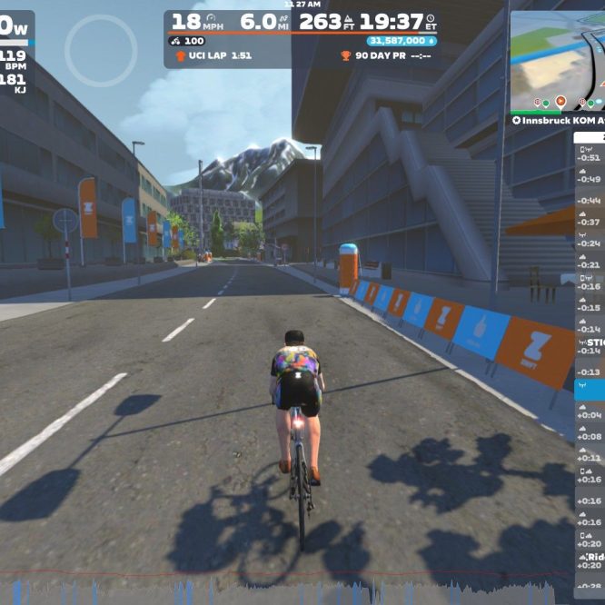 A cyclist in a virtual cycling game on Zwift rides down a digital road with buildings and trees on either side. The screen displays information about speed, distance, and heart rate, as well as a leaderboard with nearby cyclists. Colorful flags mark the path, and a mountainous landscape is visible in the background.